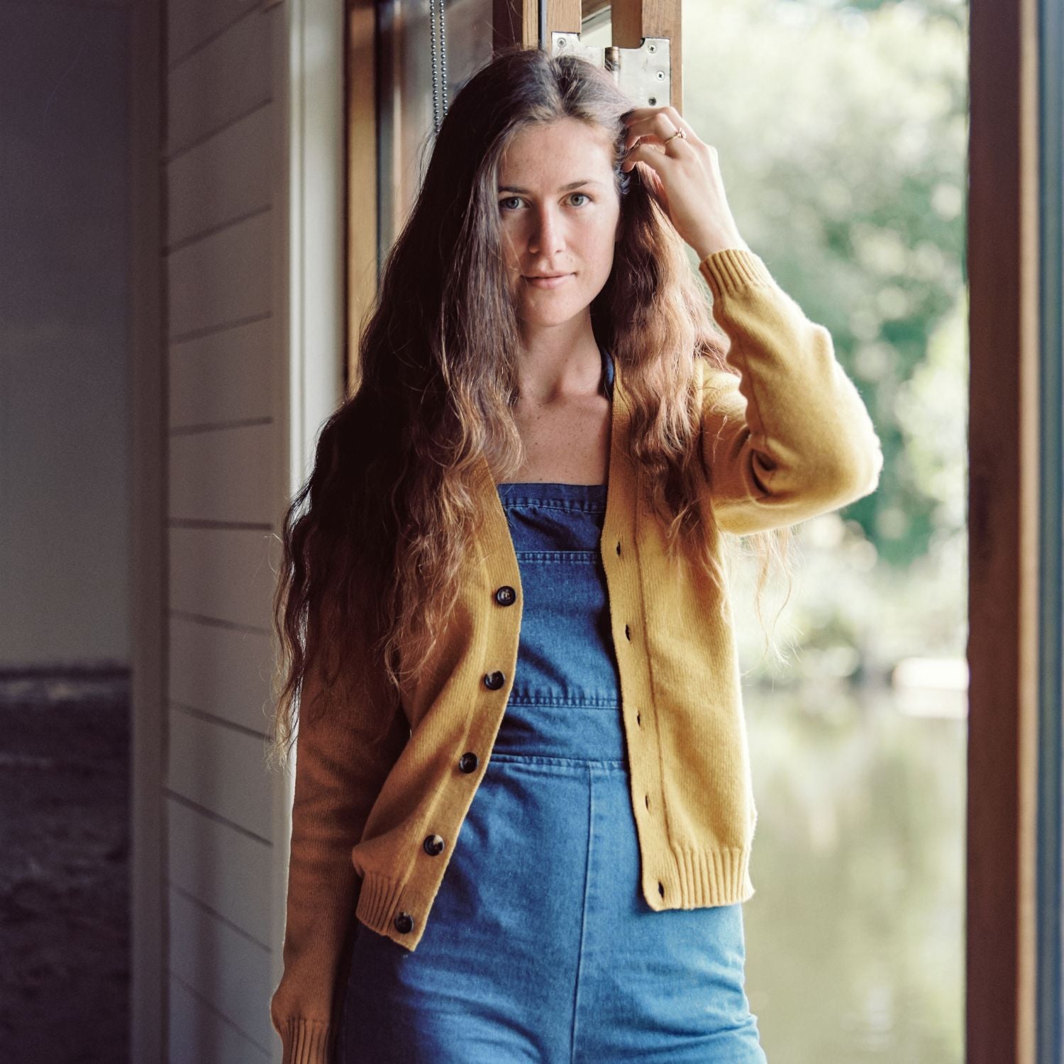 A woman standing by a window wearing a yellow cardigan over a blue dress, with her hand running through her long brown hair.