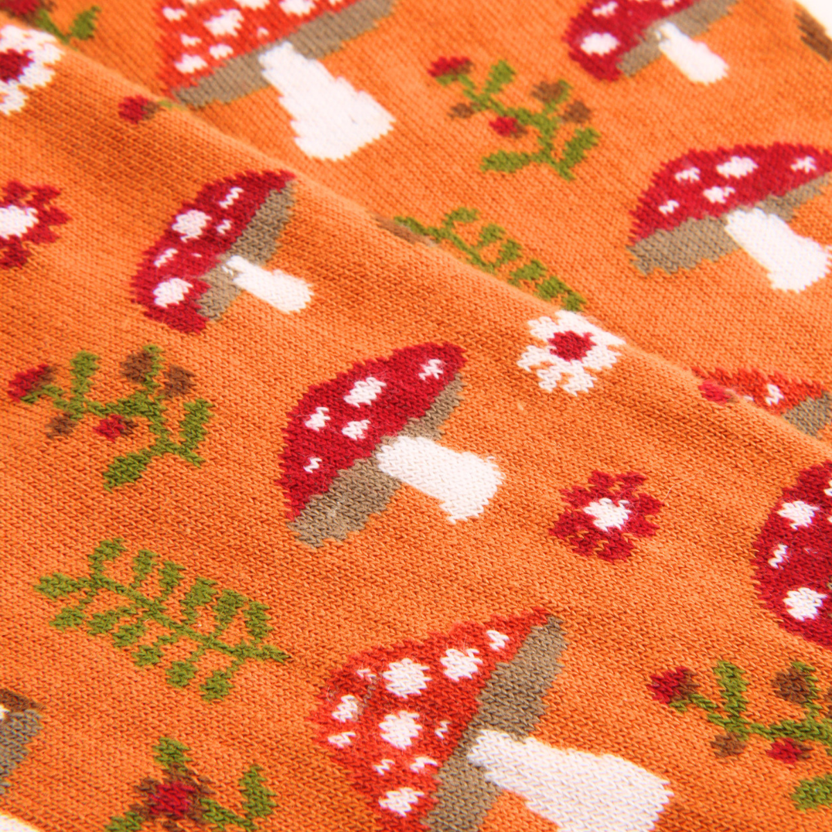 Close-up of a knitted fabric with an orange base and a repeating mushroom and floral pattern in red, white, green, and gray.