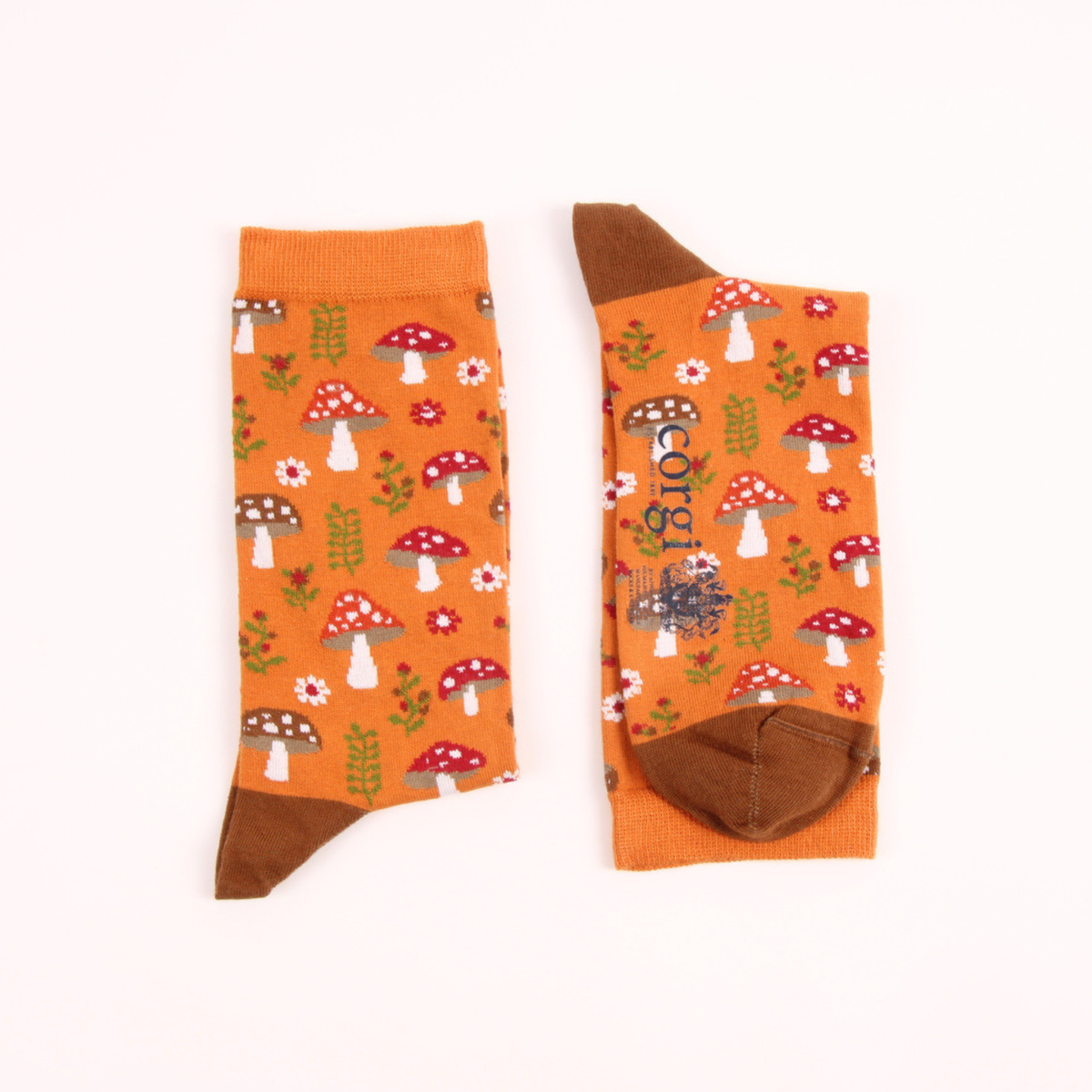 A pair of orange crew socks with a whimsical mushroom and flower pattern, featuring solid brown heels, toes, and cuffs, displayed flat against a white background.