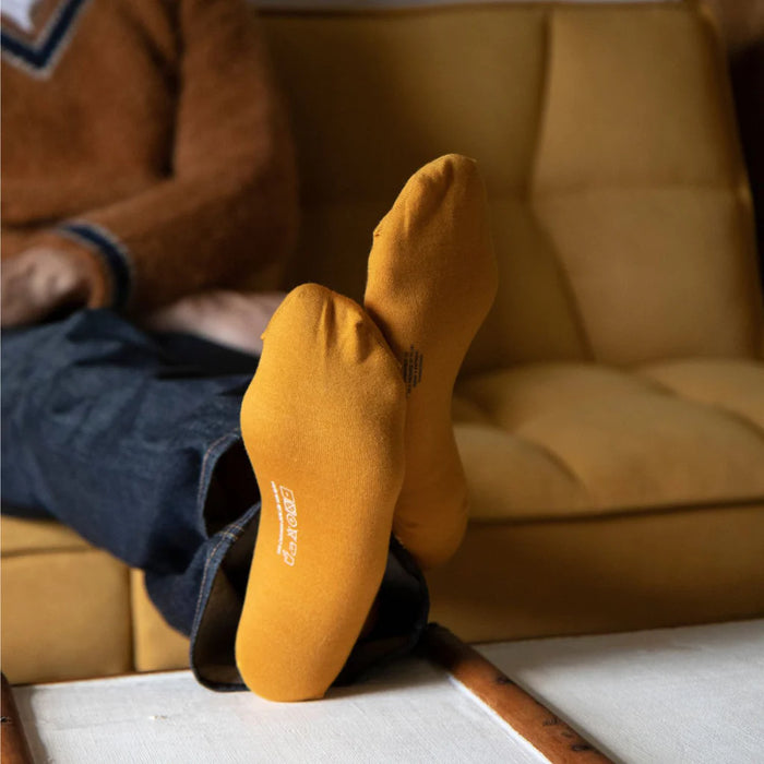 A person seated with their feet up on a table wearing mustard yellow cashmere and silk socks with a small ribbed cuff.