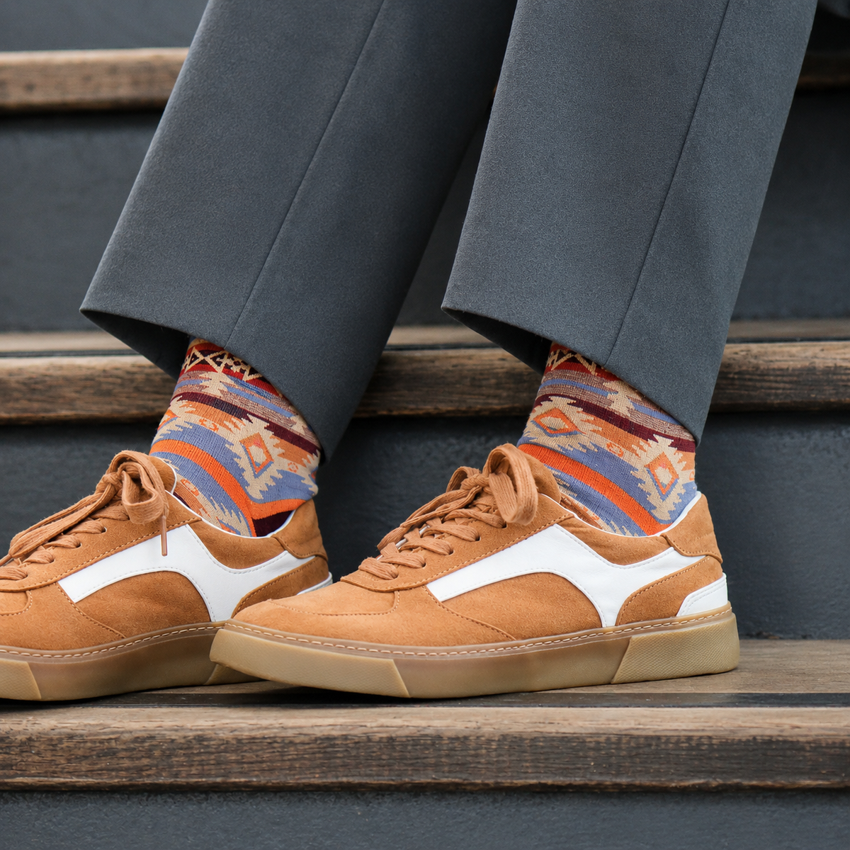A person wearing brown suede sneakers with white side stripes, gray trousers, and colorful patterned socks, sitting on wooden steps.