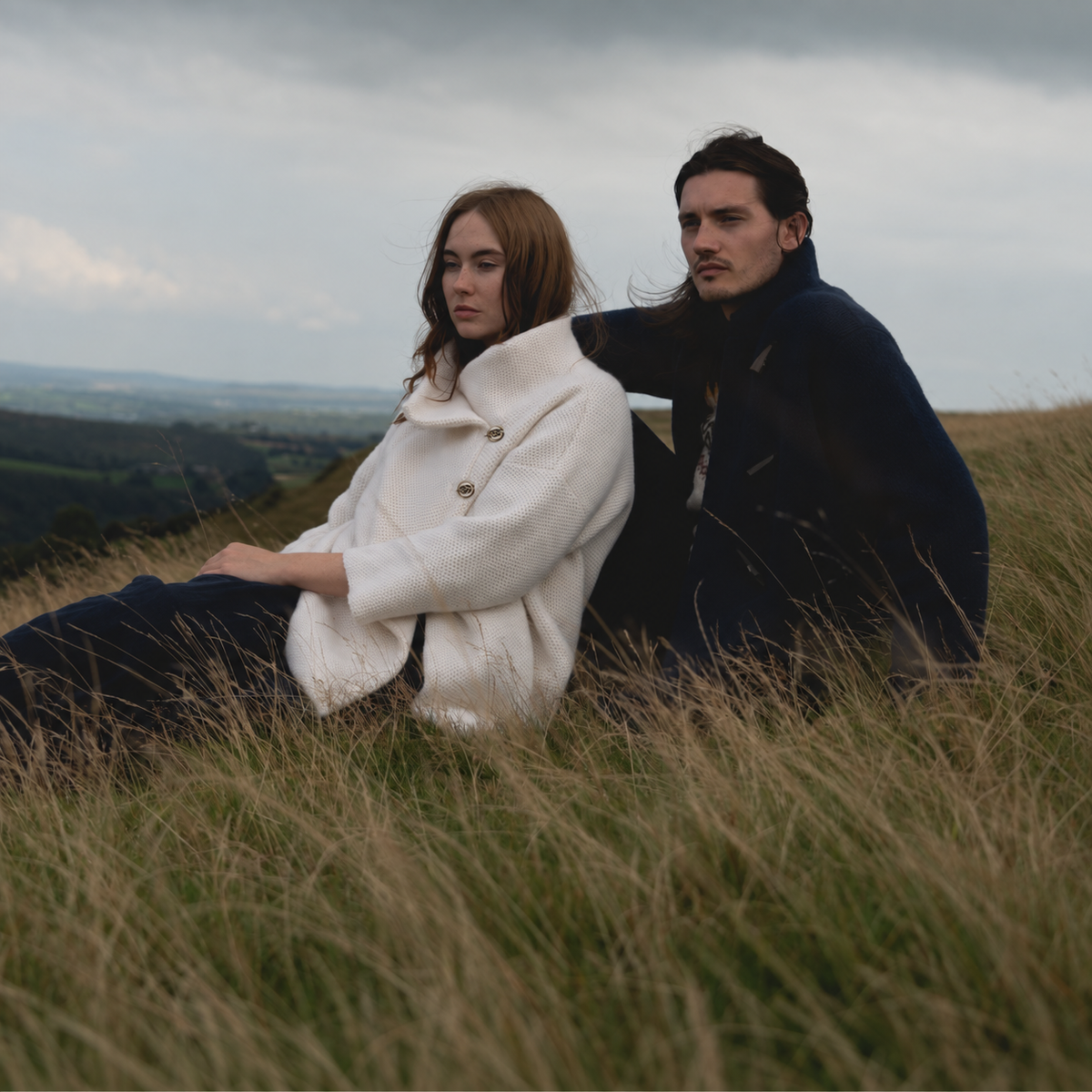 A man and a woman sit together on a grassy hillside under an overcast sky. The woman wears a cream-colored textured coat, and the man wears a dark navy coat.