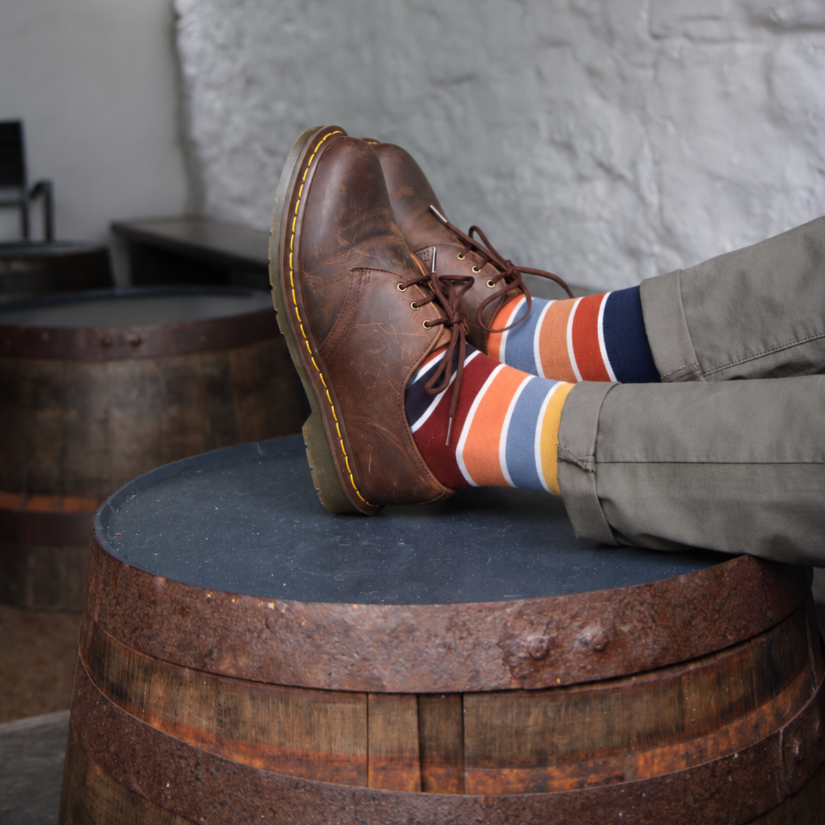 A person wearing a brown leather lace-up shoe and colorful striped socks, sitting with their legs crossed on top of a rustic wooden barrel in an indoor setting.