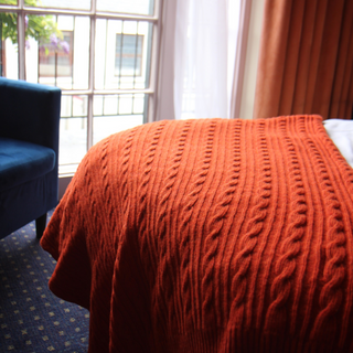 Orange knitted blanket on a bed with a blue chair and window in the background