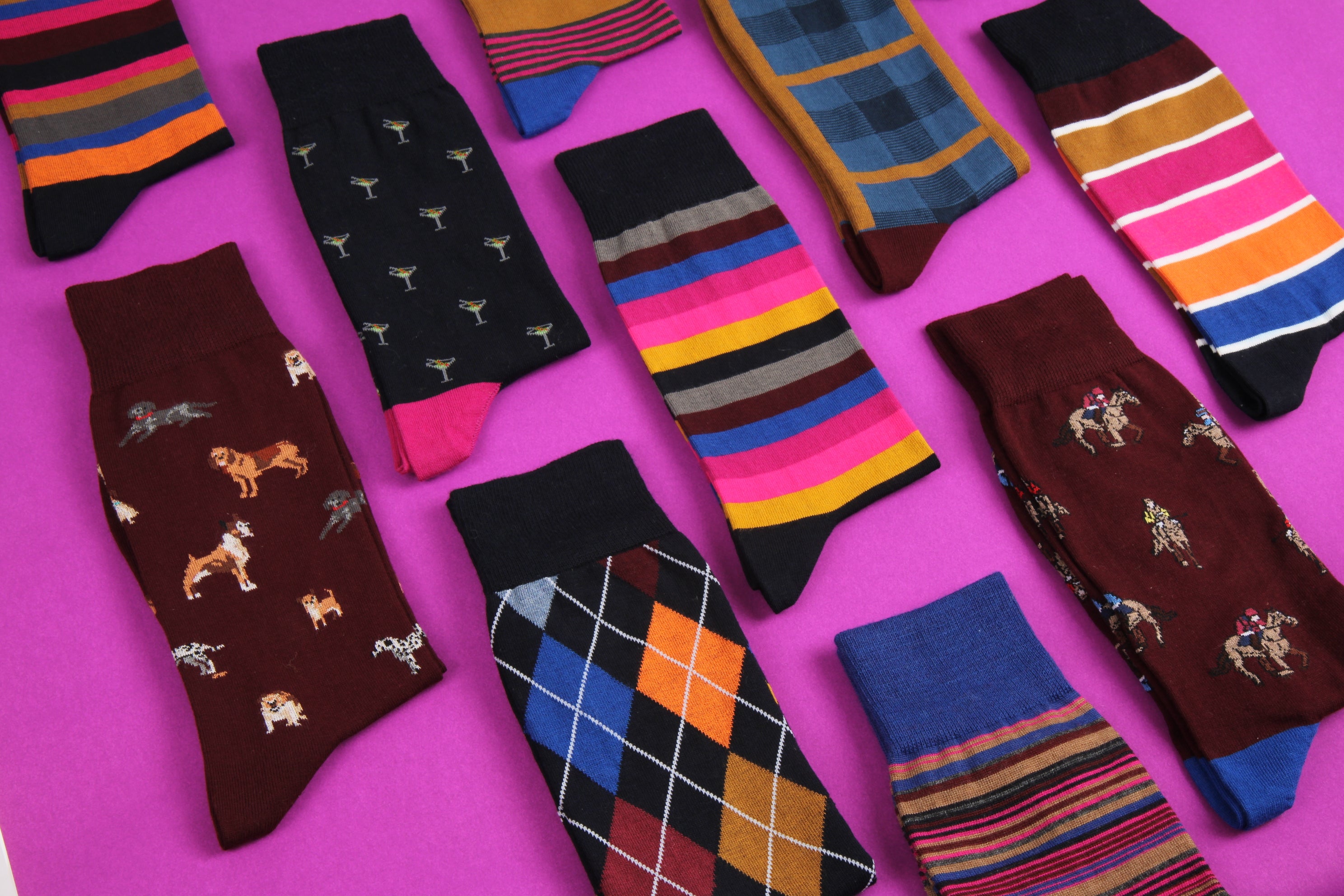 A flat-lay arrangement of ten different men's socks in various patterns (stripes, argyle, dog prints, horse prints, plaid) on a vibrant magenta background.