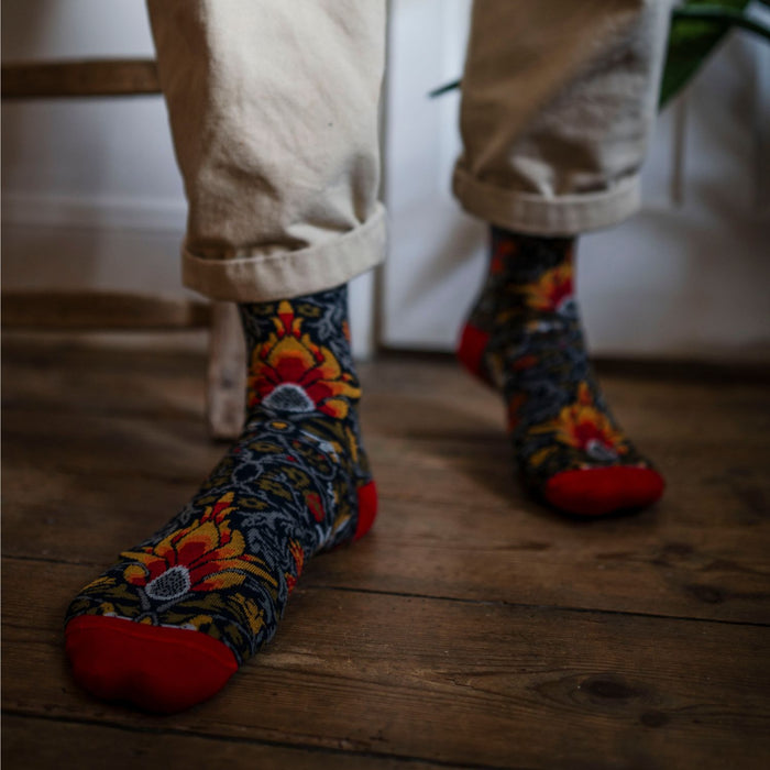 A person wearing patterned cotton socks with red tips and a floral design, standing on a wooden floor.