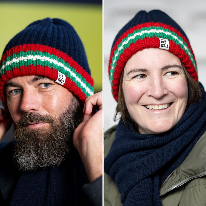 Two models wearing the navy ribbed beanie hat with red and green stripes, one male and one female, smiling towards the camera.