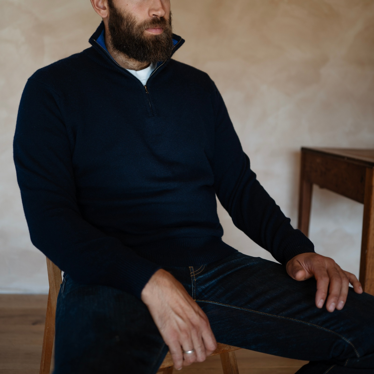 A man wearing a dark navy quarter-zip sweater and dark jeans, seated on a wooden chair in a warm, rustic interior setting.