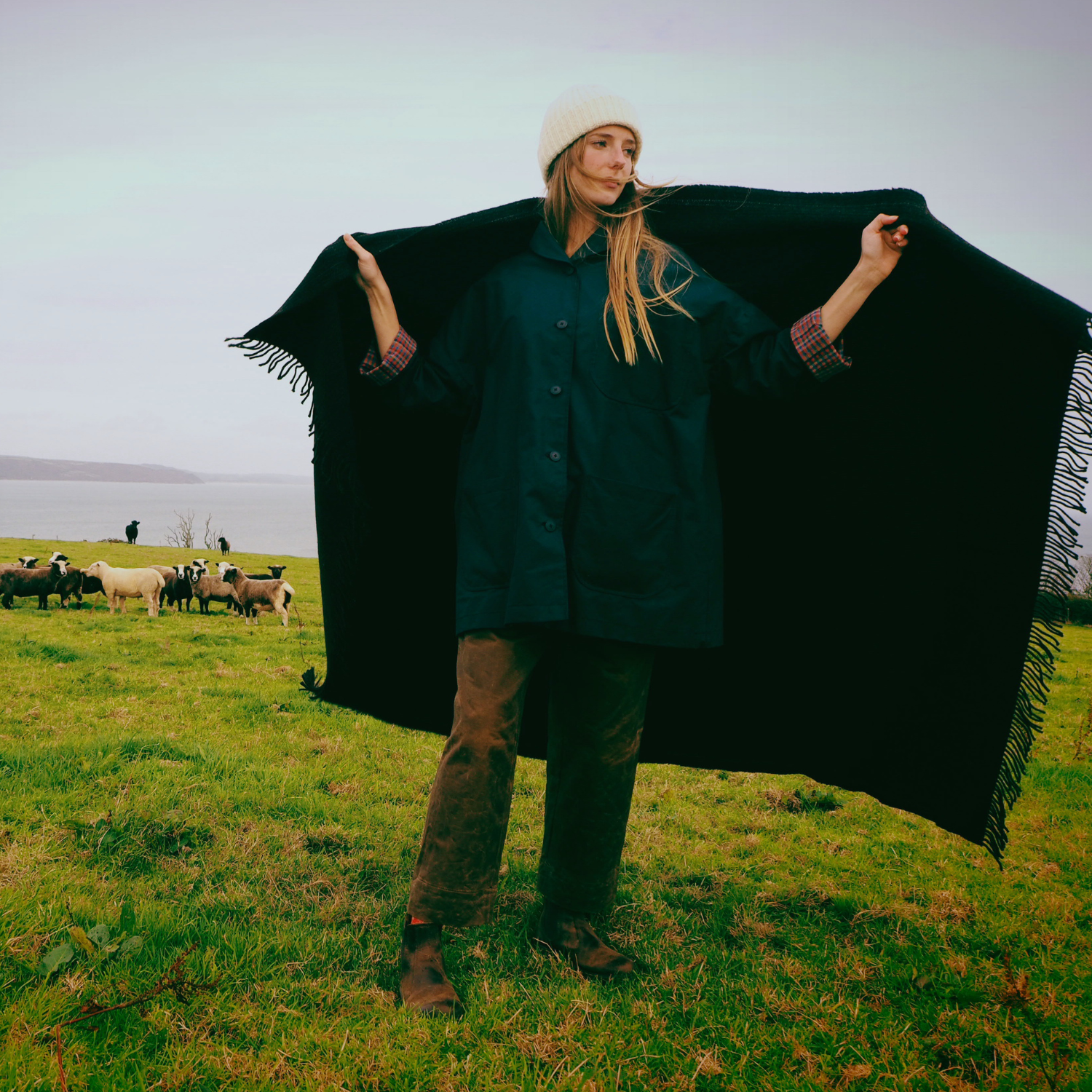 Person holding a black blanket in a grassy field with sheep in the background