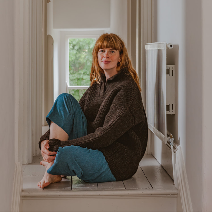 Woman sitting on a windowsill wearing a dark sweater and blue pants, with a neutral background.