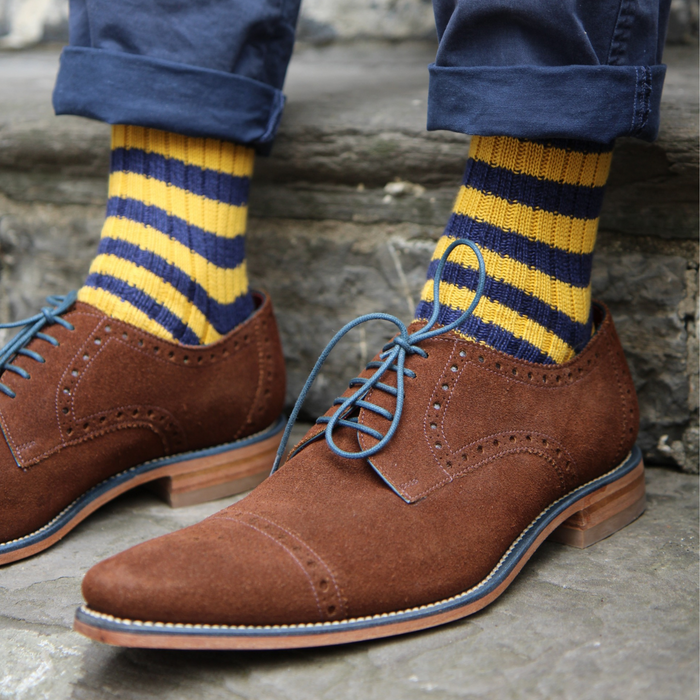 Brown suede shoes with blue laces worn with yellow and blue striped socks on a stone surface.