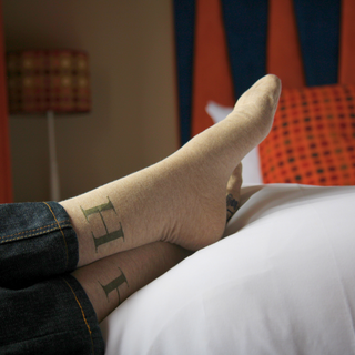Close-up of feet wearing beige socks with a 'H' letter, lying on a bed with colourful bedding in the background.
