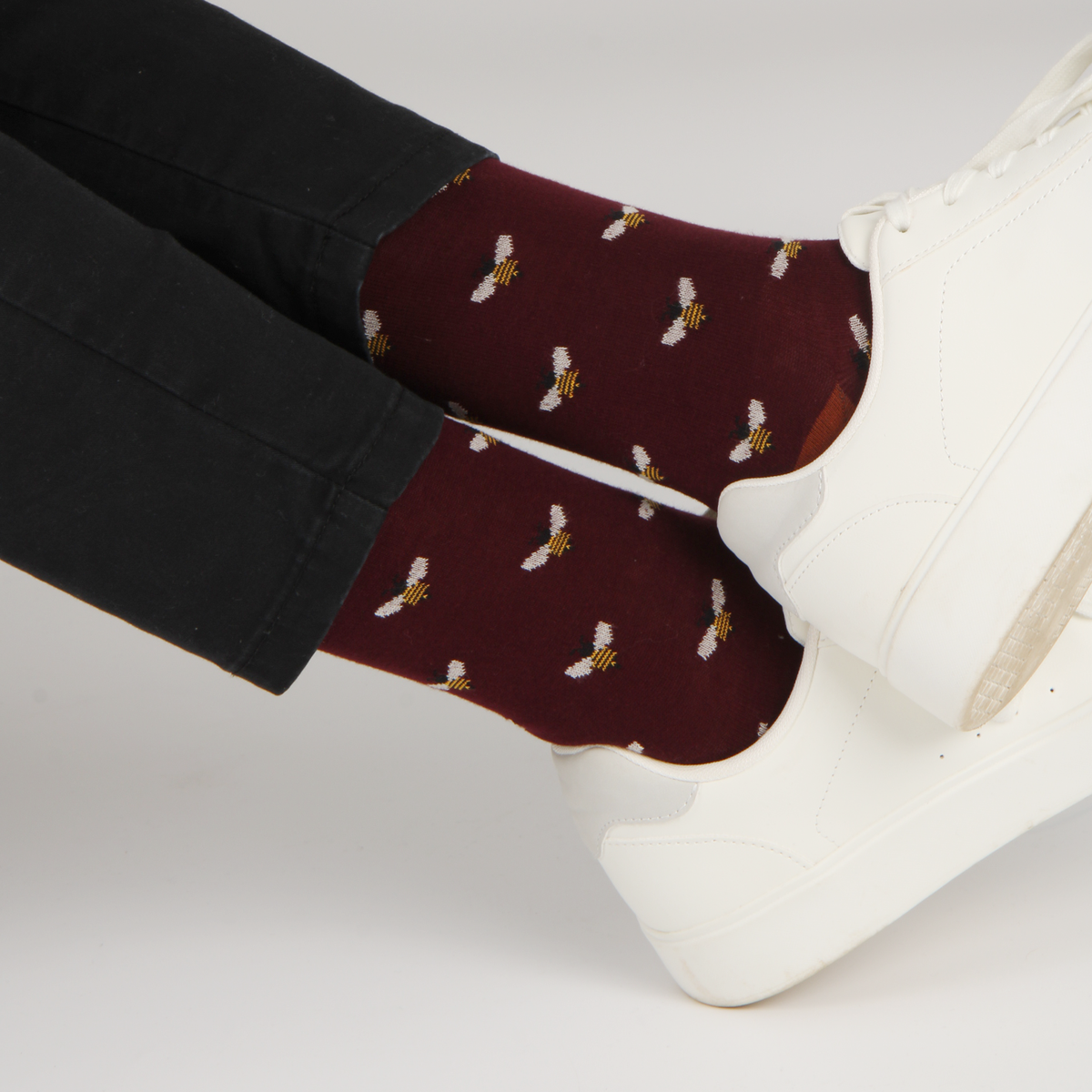 A close-up of a person's feet wearing dark burgundy crew socks with a repeating pattern of small white bees with yellow accents, paired with black pants and white platform sneakers.