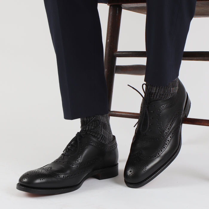A pair of men's formal woven check cotton socks in black and white, displayed alongside a pair of black dress shoes and navy pants.