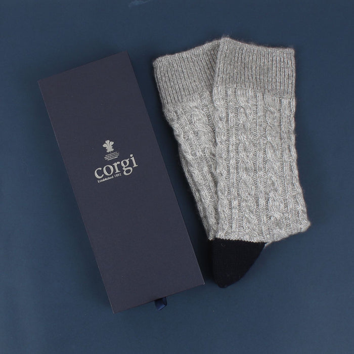 A pair of hand-knitted gray cable cashmere and cotton socks displayed alongside its packaging box with the brand 'Corgi' visible.