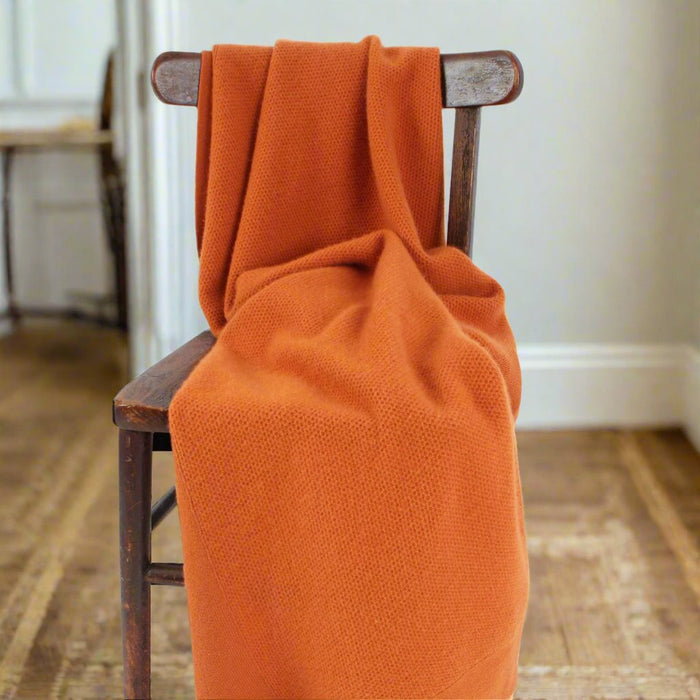 An orange wool blanket with a fine honeycomb texture draped over the back of a wooden chair, set against a background of a simple indoor scene with a dark chair and a white door.