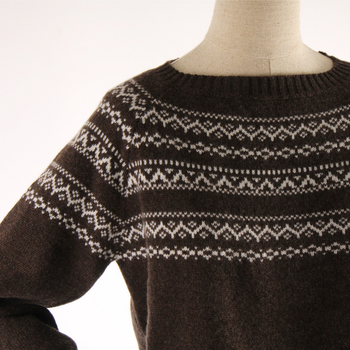 A close-up of a dark brown knit sweater with a cream-colored Fair Isle pattern across the yoke and sleeve, displayed on a mannequin torso against a white background.