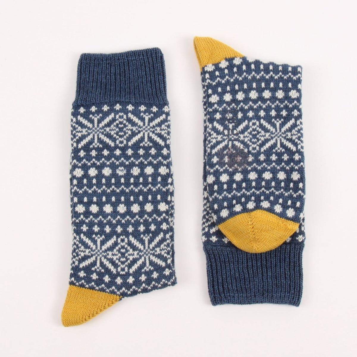 A pair of navy blue knit socks with white Nordic snowflake patterns and mustard yellow heels, toes, and cuffs, displayed flat on a white background.