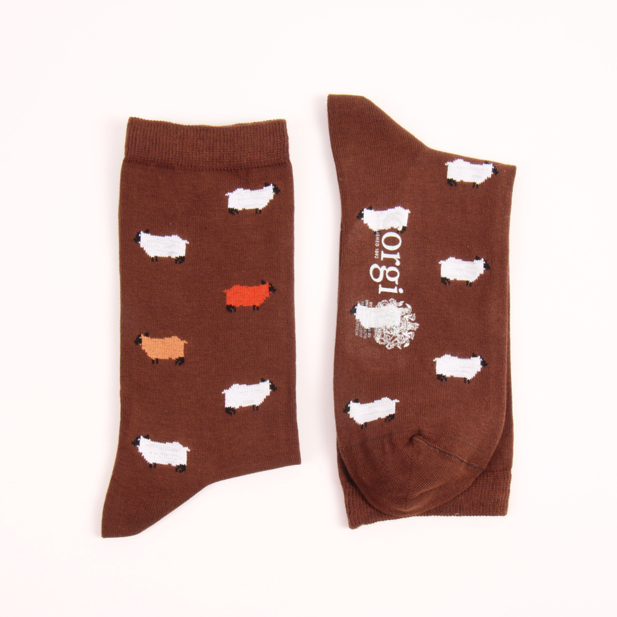 A pair of brown crew socks laid flat on a white background. One sock shows a pattern of small sheep in white, light orange, and red, while the other shows the brand name 'origi' printed vertically in white.