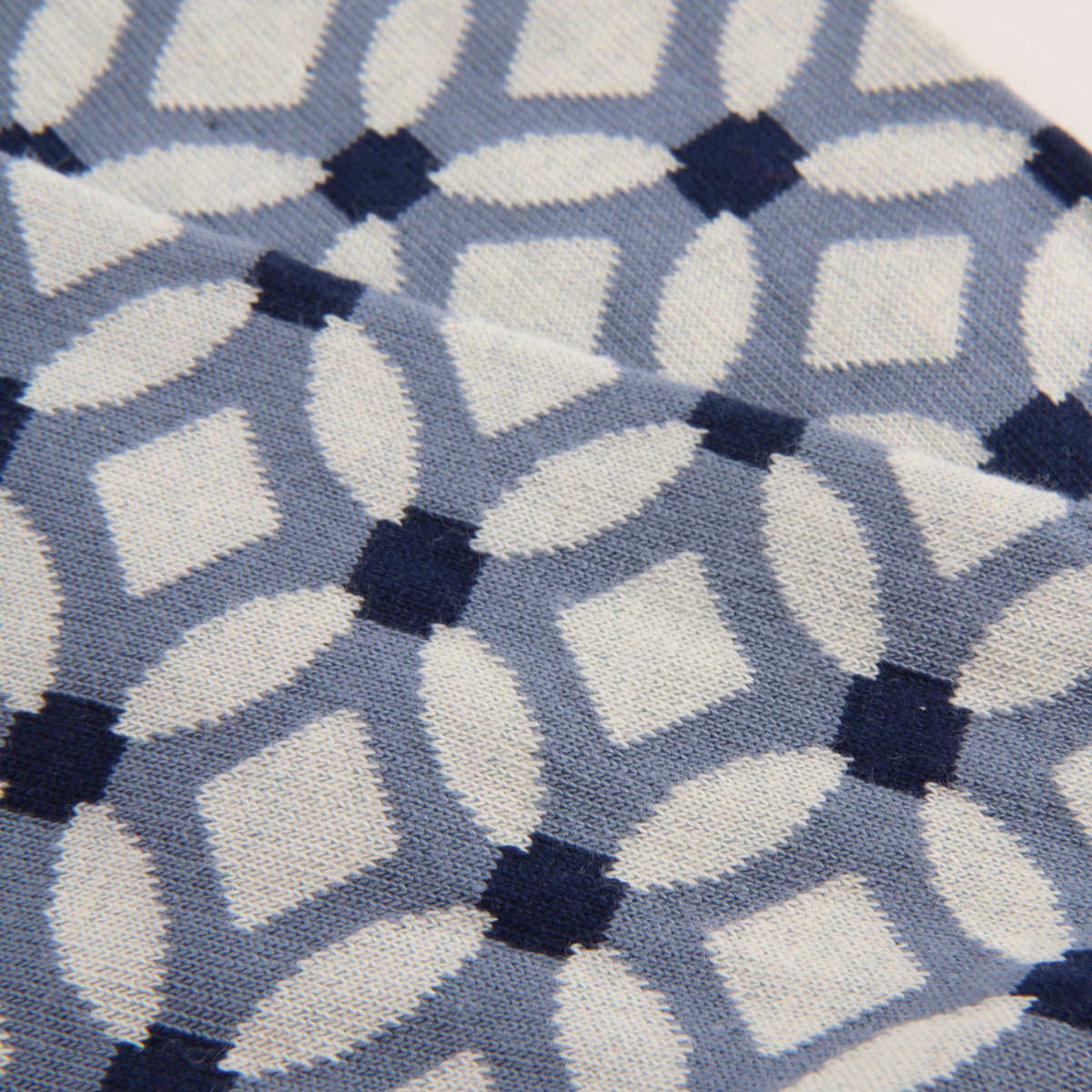 A close-up of a knit fabric with a repeating geometric pattern of white and light blue interlocking shapes, accented by dark navy blue squares.