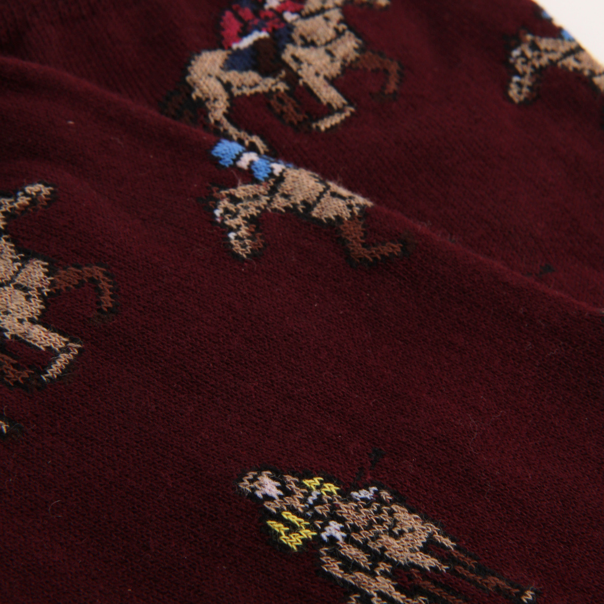 Close-up of a dark burgundy knit fabric with a repeating embroidered polo player and horse motif in beige, brown, blue, red, and yellow.