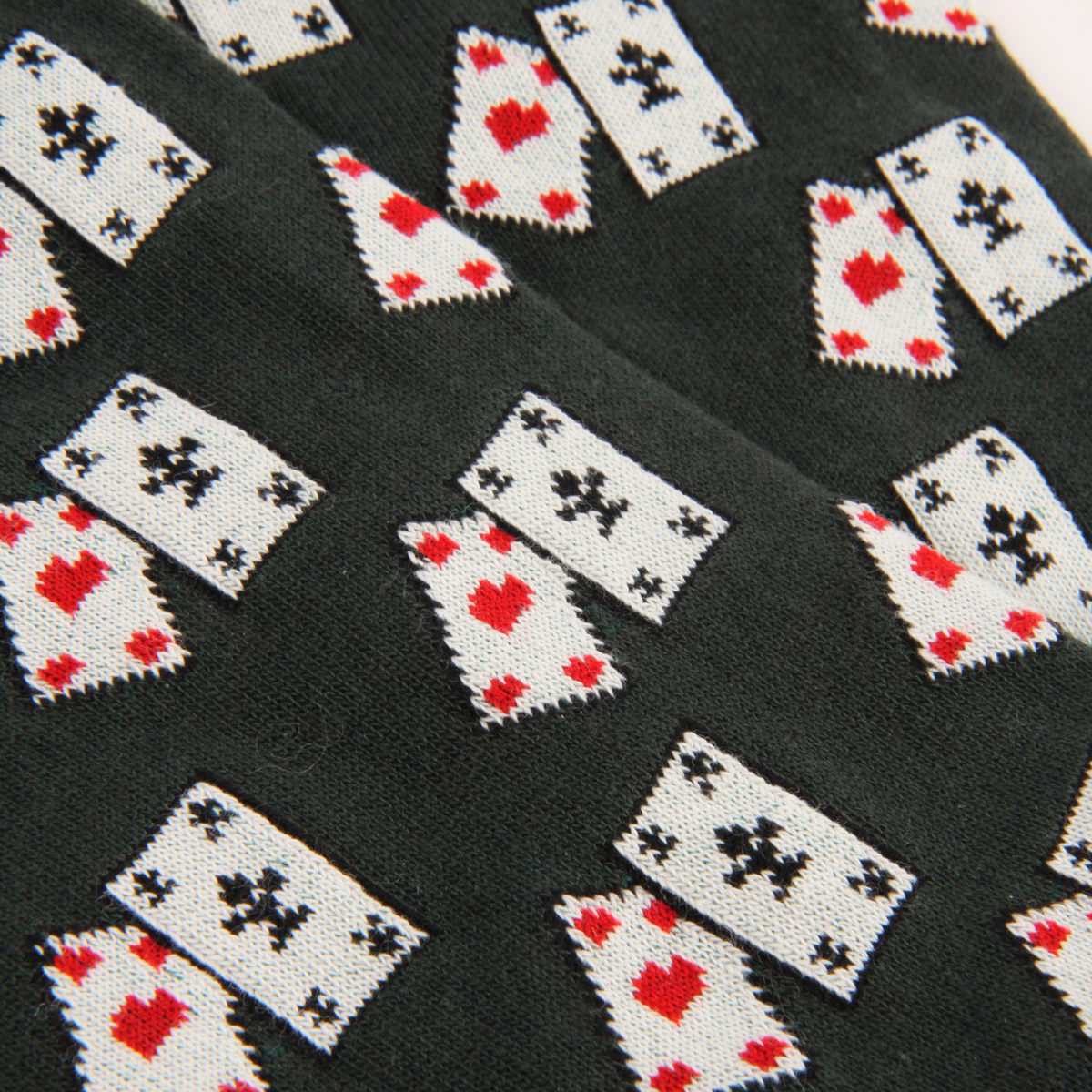 Close-up of a knit fabric with a repeating pattern of playing card suits: white cards with red hearts and black clubs on a very dark green background.