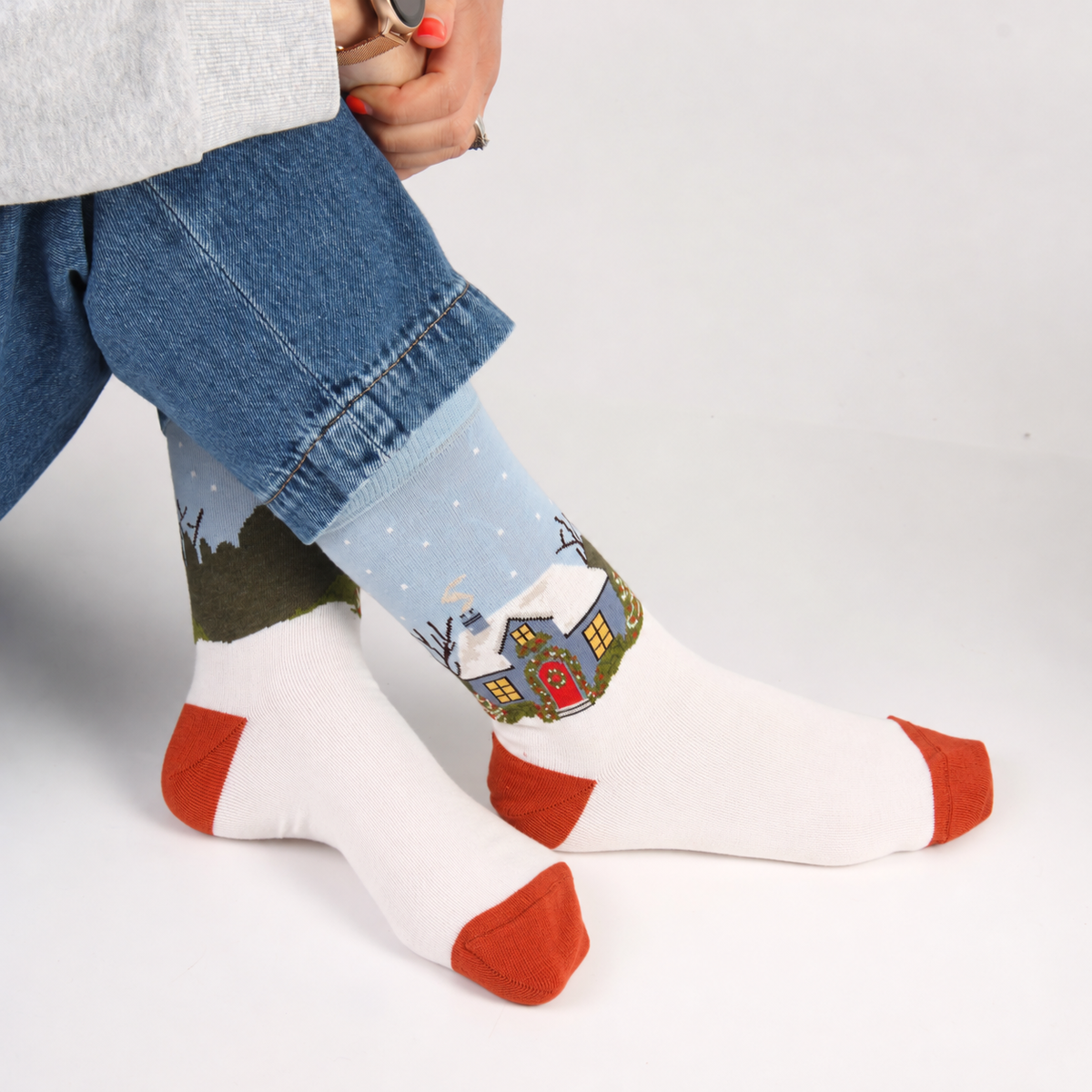 A person wearing white crew socks with rust-orange toes and heels, featuring a light blue winter village print around the cuffs. The person is also wearing blue jeans, shown from the waist down against a clean white studio background.
