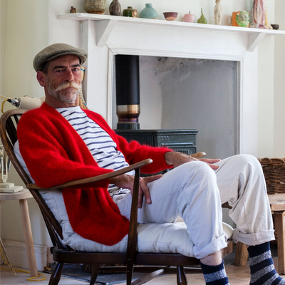 Man sitting in a chair wearing a red cardigan and white pants in a room with a fireplace and shelves.