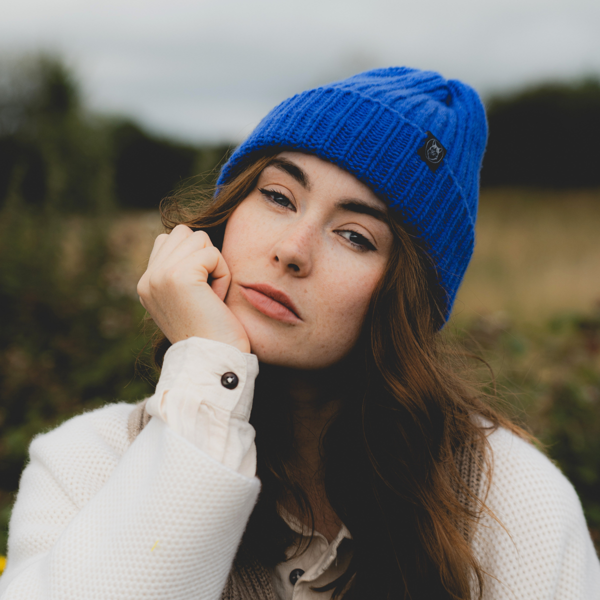 A woman wearing a bright blue ribbed knit beanie with a small black button logo, posed outdoors against a soft-focus natural background.