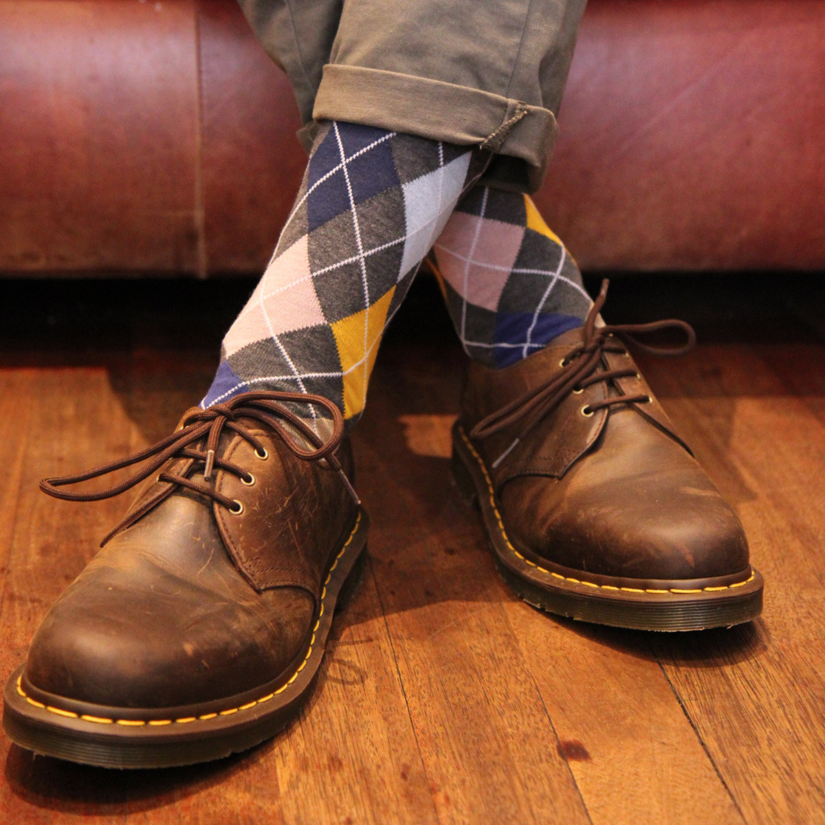 A close-up of a person's legs wearing brown leather Dr. Martens shoes and argyle socks in navy, yellow, pink, and white, sitting on a brown leather couch over a wooden floor.