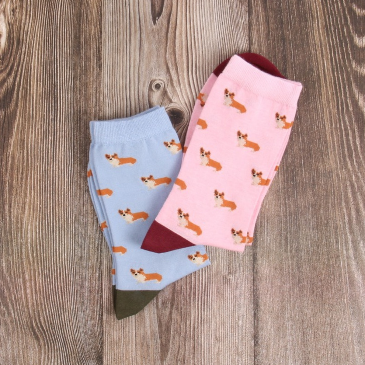 Two pairs of patterned socks laid flat on a rustic wood surface. One pair is light blue with an orange and cream corgi print and olive green toes.