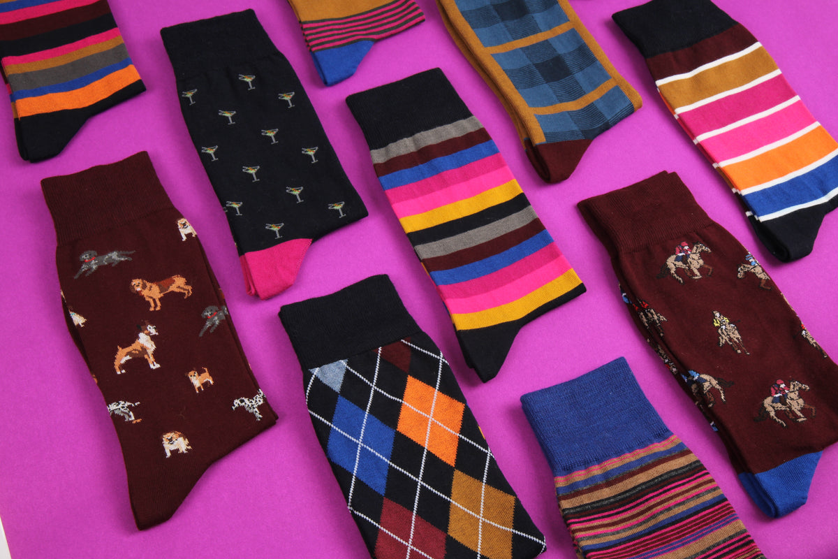 A flat-lay arrangement of ten different men's socks in various patterns (stripes, argyle, dog prints, horse prints, plaid) on a vibrant magenta background.