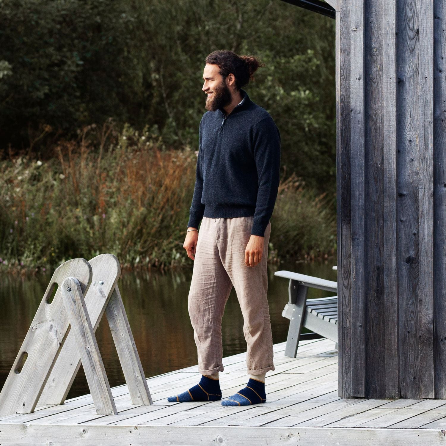 A man wearing a half zip sweater with a contrasting color collar and a leather puller. The sweater is predominantly grey, and the man is standing outdoors next to a body of water and a wooden structure.