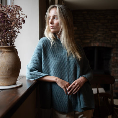 A woman wearing a blue textured cashmere poncho standing near a fireplace, with a vase of flowers beside her.