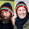 Two models wearing the navy ribbed beanie hat with red and green stripes, one male and one female, smiling towards the camera.
