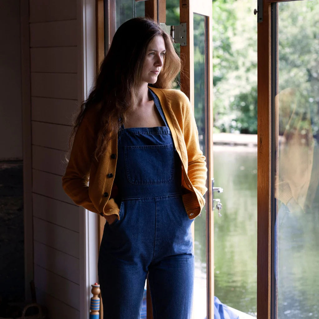 Woman in blue overalls and yellow cardigan standing by a door with a view of water and trees.