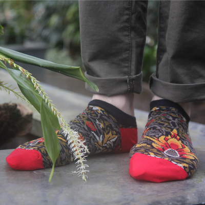 A pair of multicolored trainer socks with a geometric pattern, predominantly red and black, worn on someone's feet, with greenery in the background.