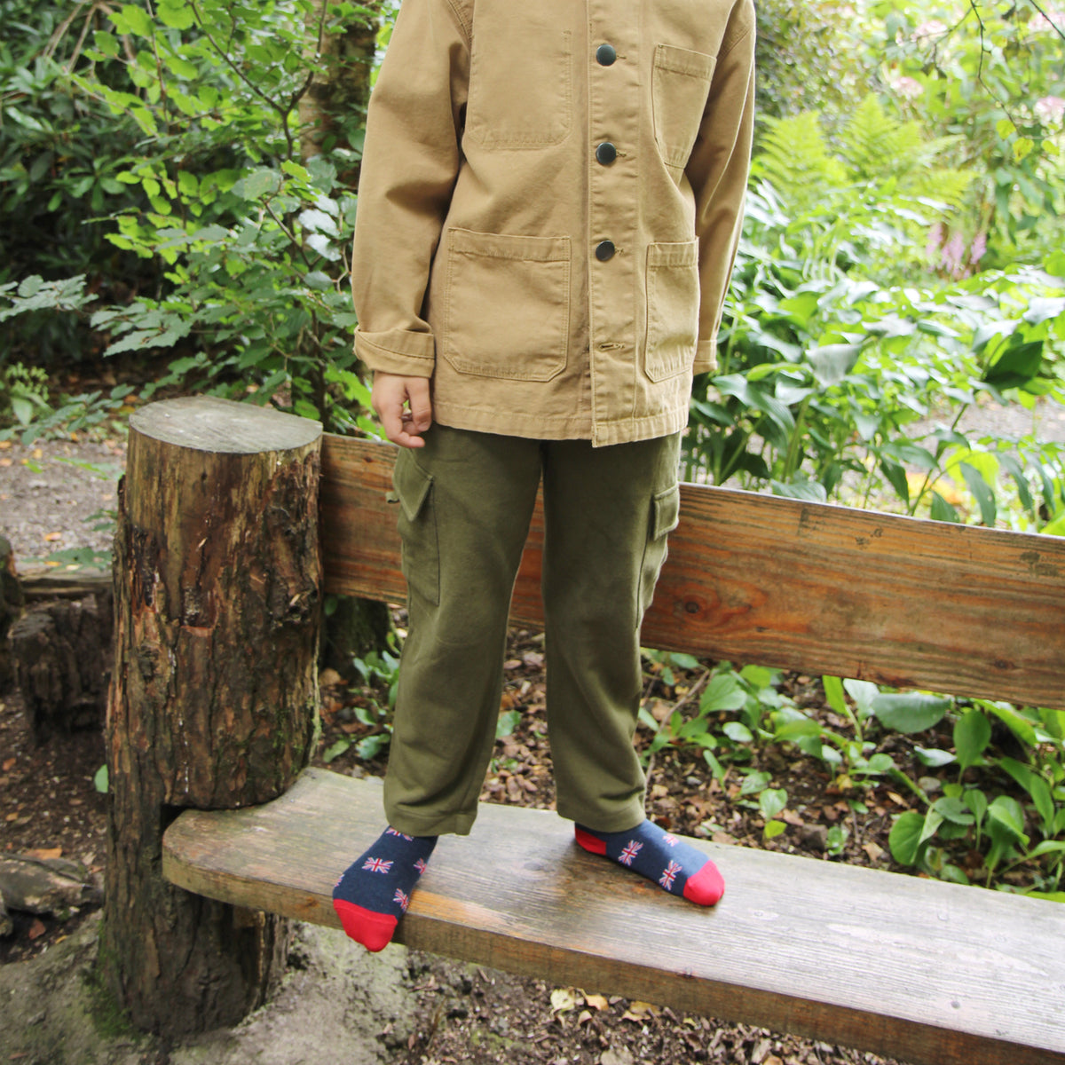 Child wearing a beige jacket and green pants sitting on a wooden bench in a forest setting.