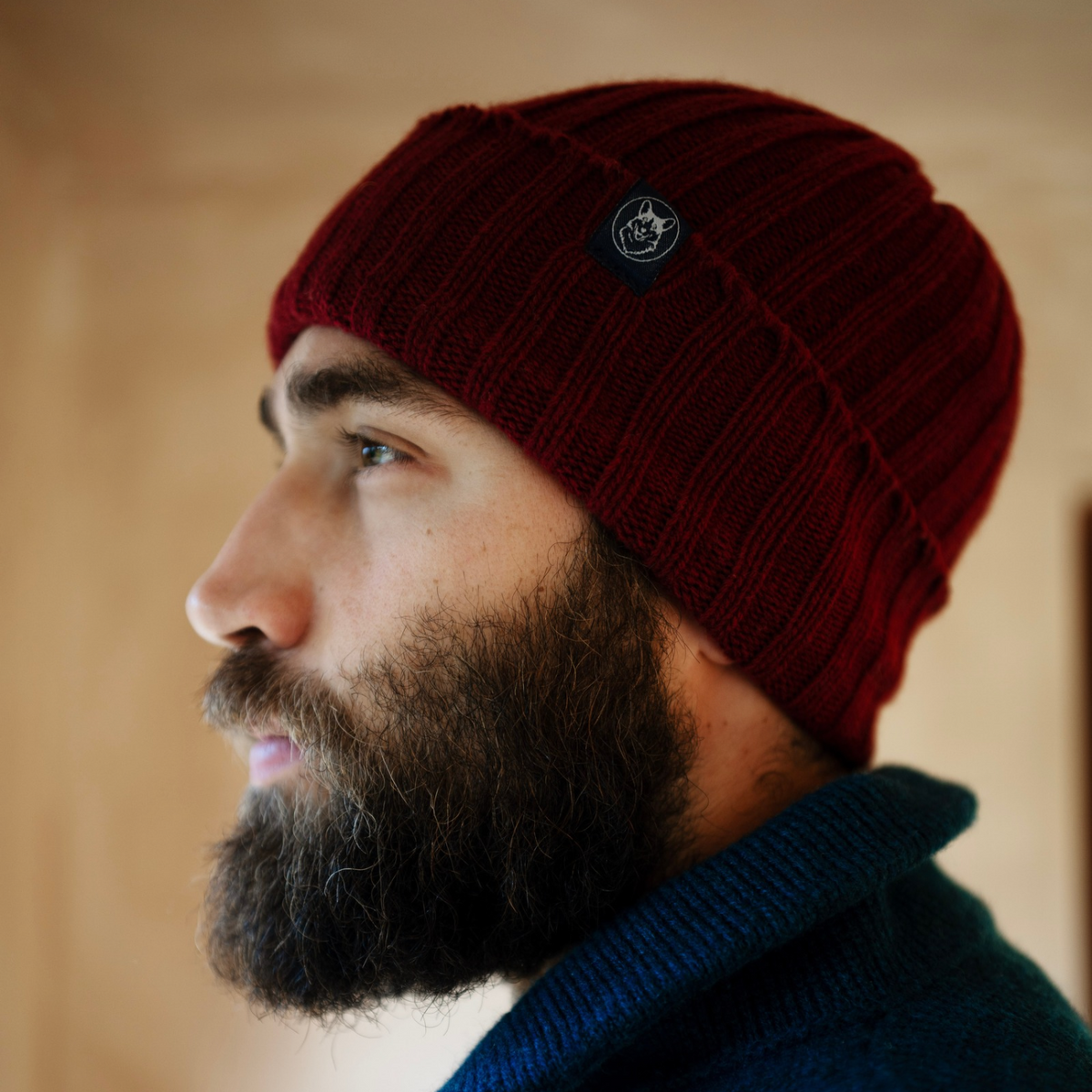 A close-up side profile of a man with a beard wearing a dark red ribbed knit beanie with a small circular logo patch and a dark blue knit sweater, set against a warm beige background.