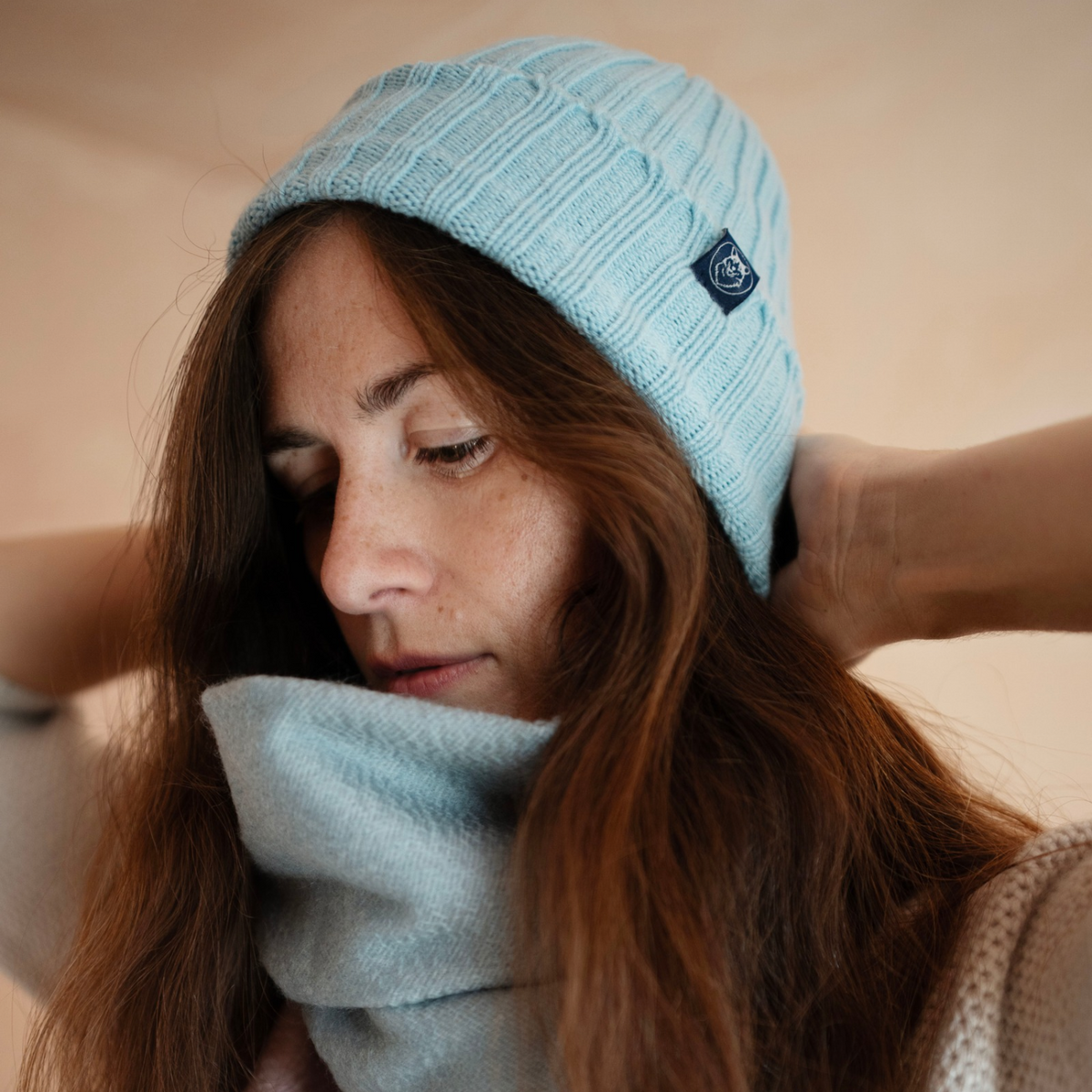 A woman wearing a light blue cable-knit beanie and a matching light blue knit neck warmer, with her hands adjusting the beanie against a soft beige background.