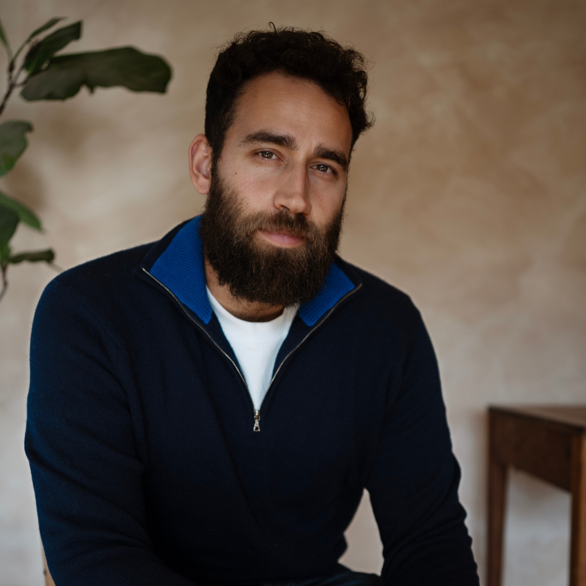 A man with a dark beard and curly hair wearing a navy blue quarter-zip sweater with a bright blue collar over a white t-shirt, sitting in a warm indoor setting.