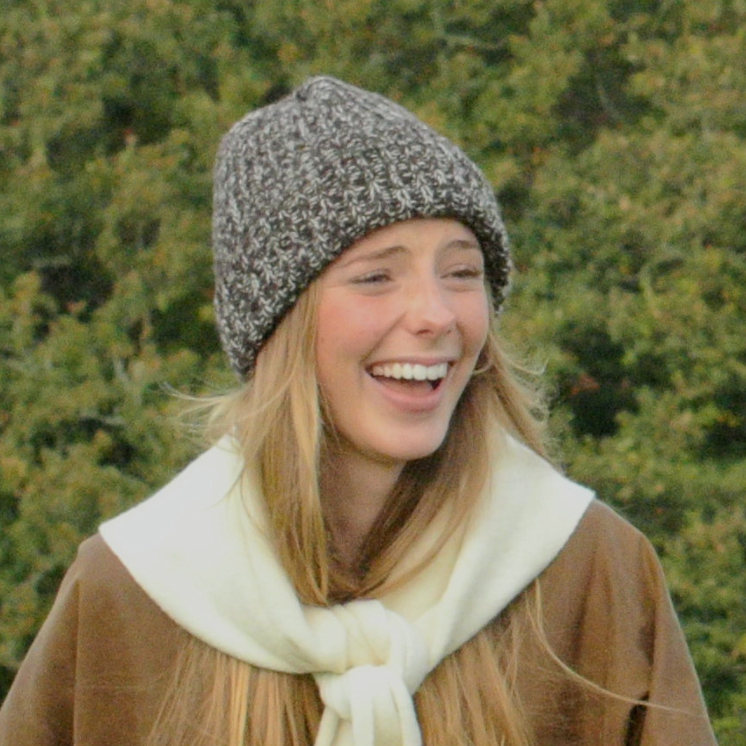 Woman wearing a knitted hat and scarf outdoors with greenery in the background
