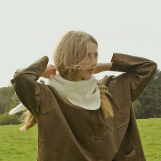 Woman in a brown coat standing in a grassy field with trees in the background wearing a white triangular scarf