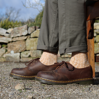 Brown leather shoes worn with rolled-up green pants and patterned socks, standing on a stone path.