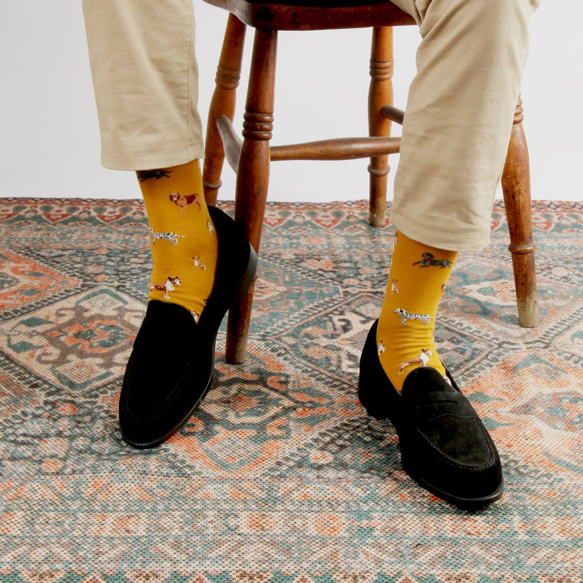 A person seated on a wooden stool wearing bright yellow patterned crew socks with small illustrated figures, black loafers, and light beige trousers, standing on a patterned rug.