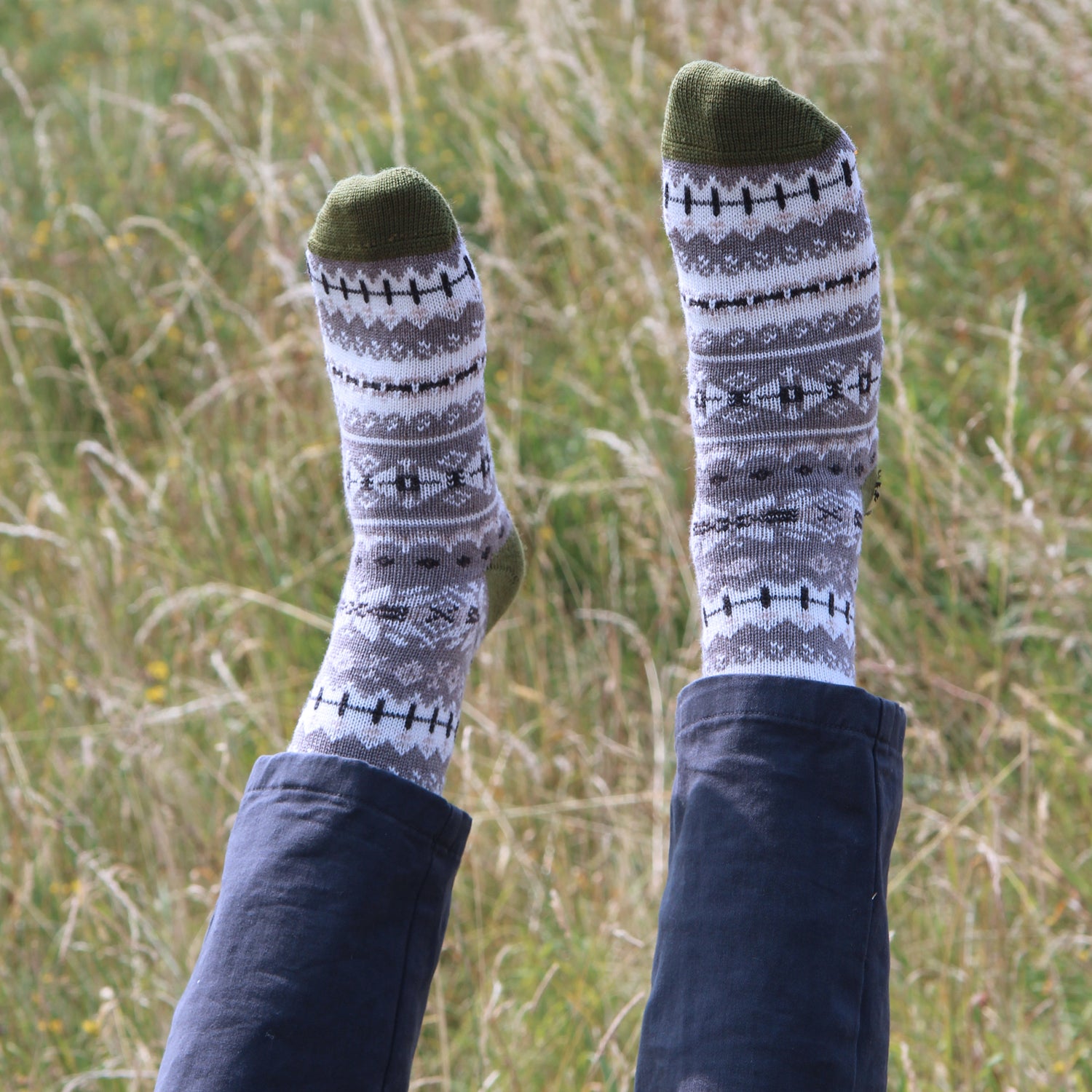 A person lying on grass outdoors with their legs raised, wearing a pair of gray and white patterned knit socks with olive green toes and heels, paired with dark blue jeans.