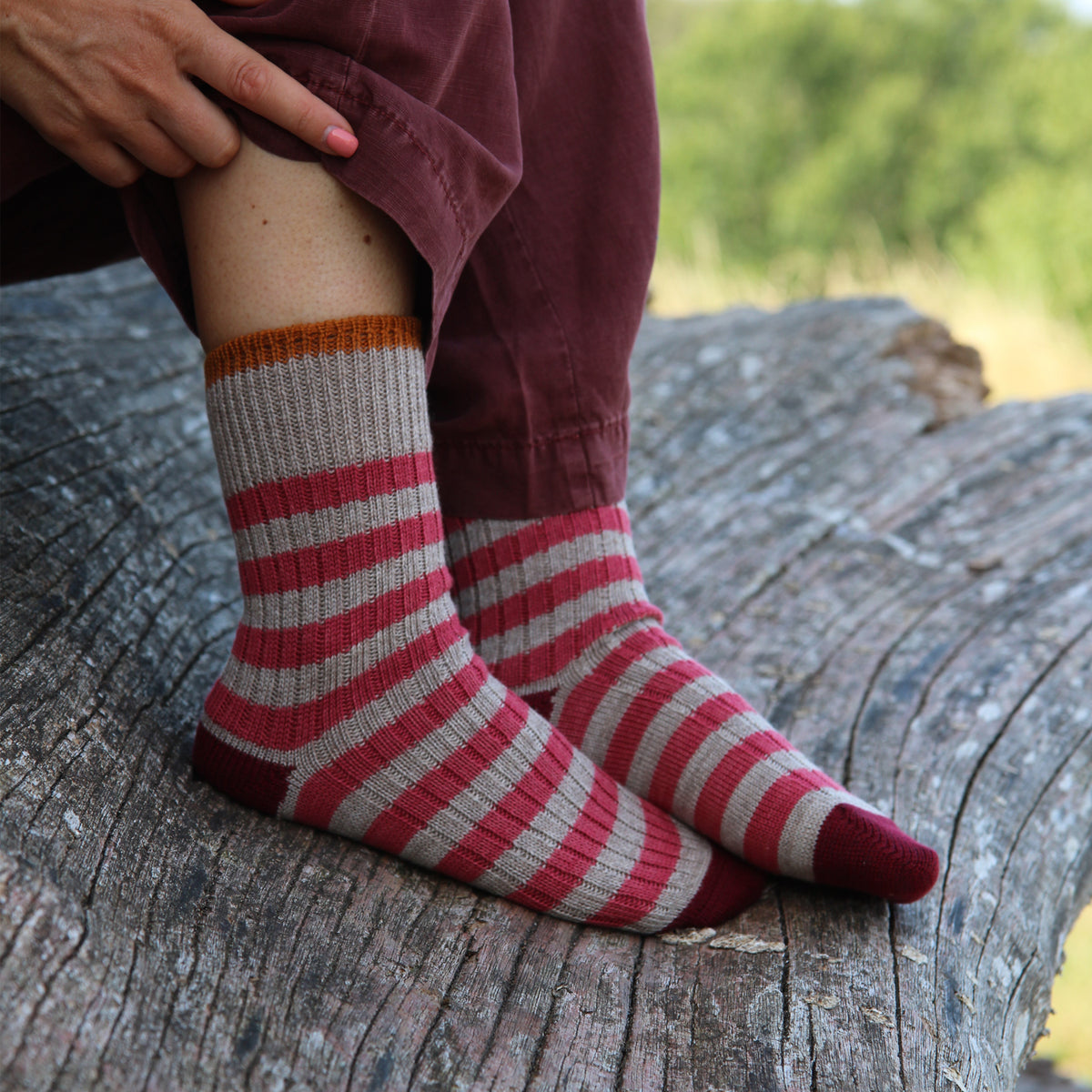 A person sitting outdoors on a weathered log wearing striped crew socks with red, dusty rose, and beige horizontal stripes, paired with burgundy pants.