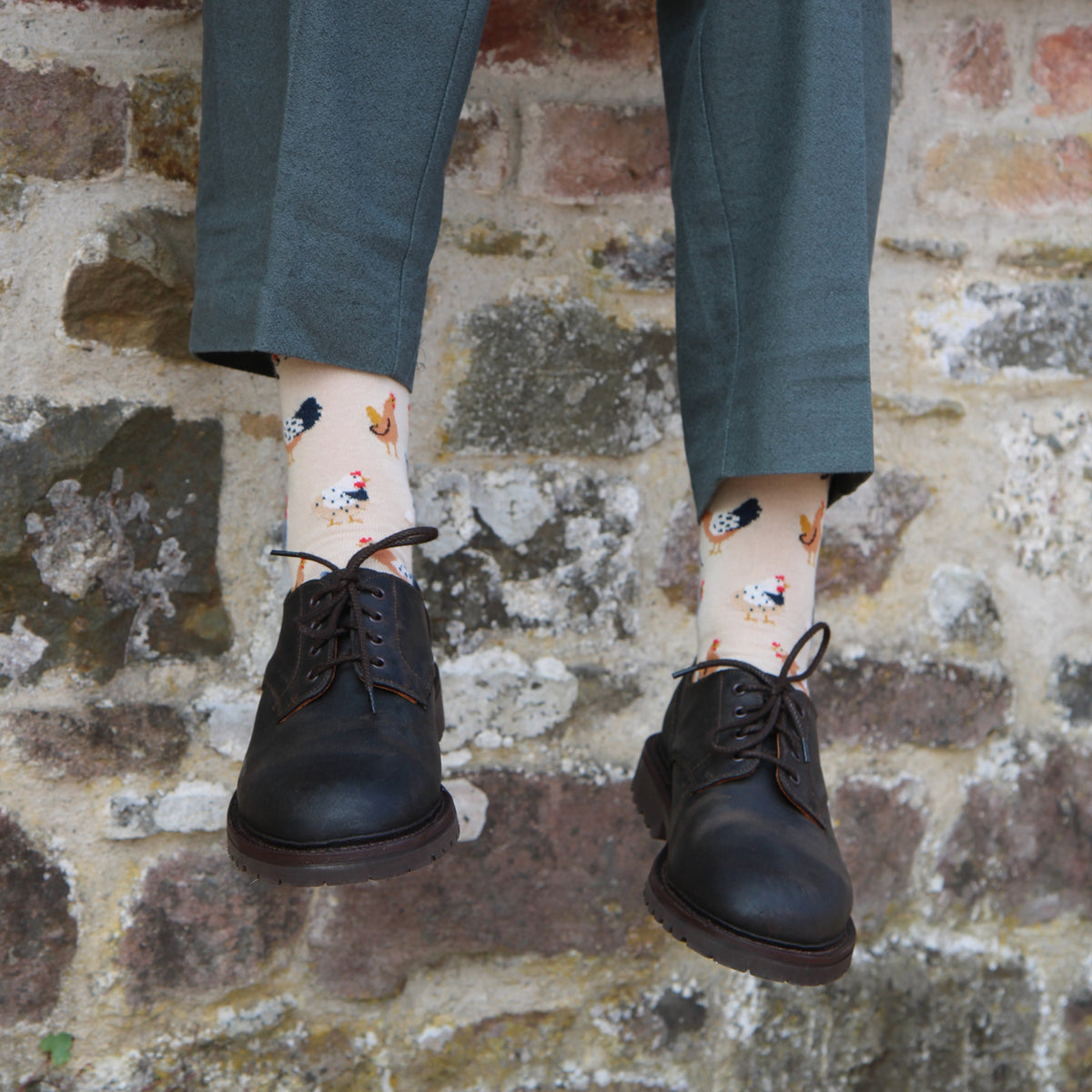 A person wearing dark gray trousers, black leather lace-up shoes, and cream-colored socks with a colorful chicken pattern, standing against a rustic brick wall.