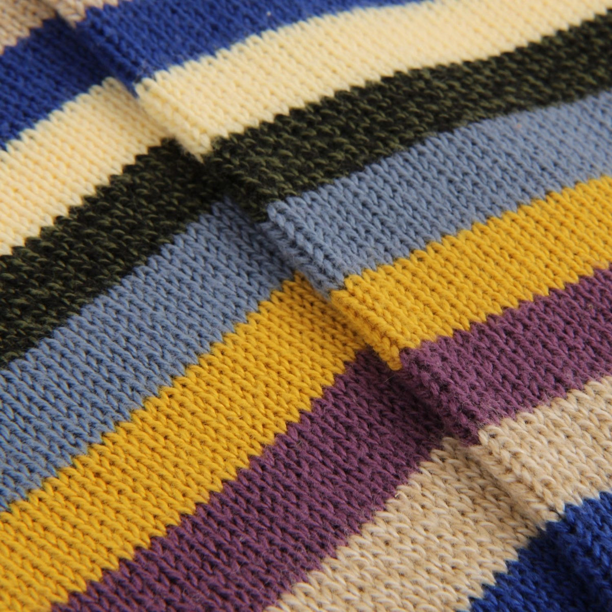 Close-up of a knitted fabric with horizontal stripes in blue, cream, dark green, light blue, mustard yellow, and purple.