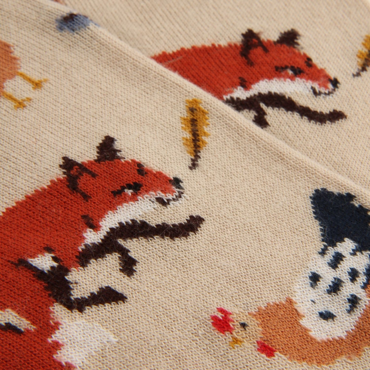 Close-up of a knitted textile with a repeating fox and bird pattern on a warm beige background.
